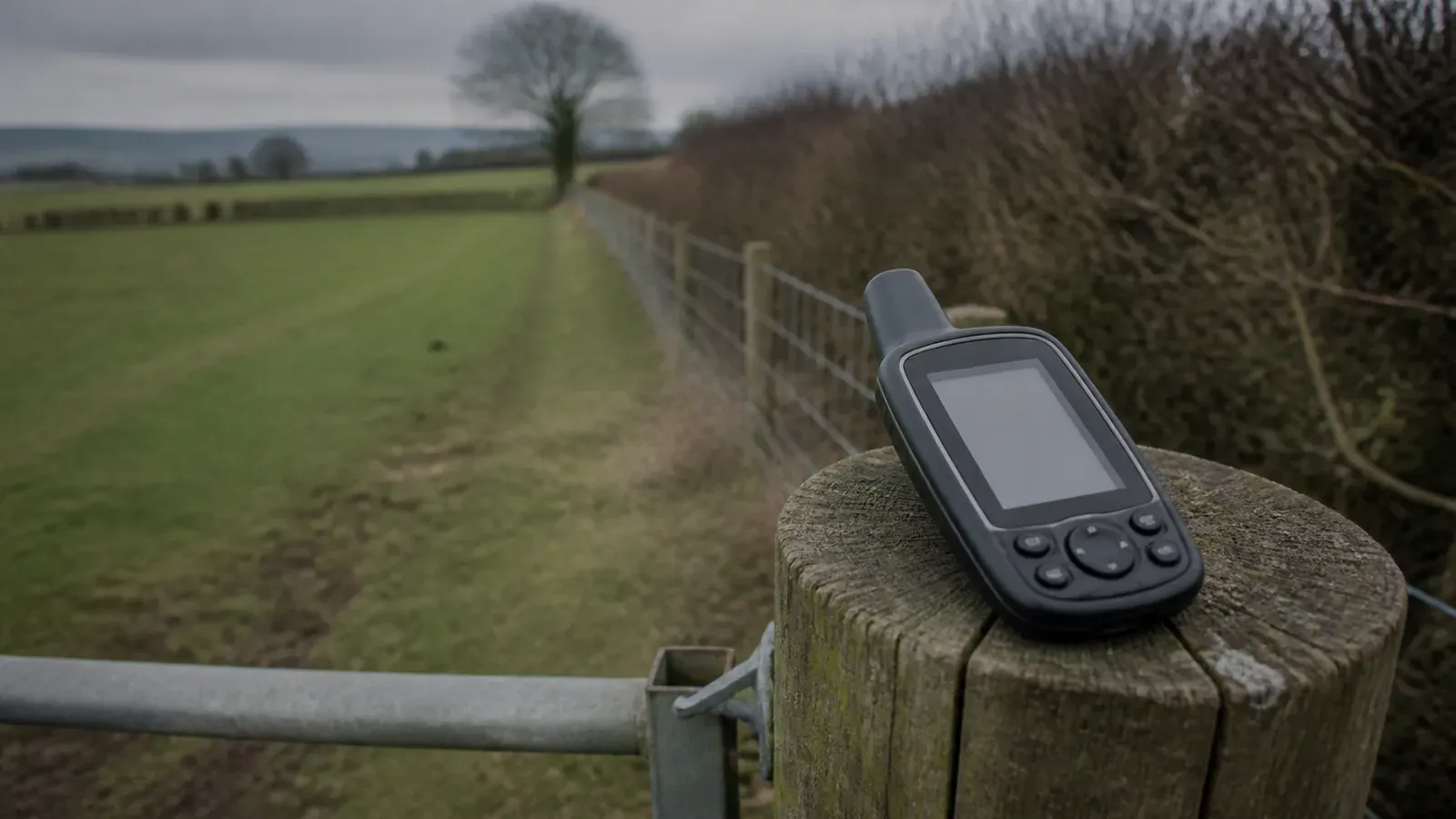 A handheld GPS receiver resting on a wooden gatepost at the corner of a smallholding, with the mapped field boundary running away behind it